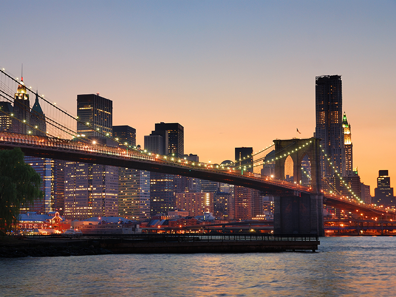 New York City Brooklyn Bridge and Manhattan skyline with skyscrapers over Hudson River illuminated with lights at dusk after sunset.